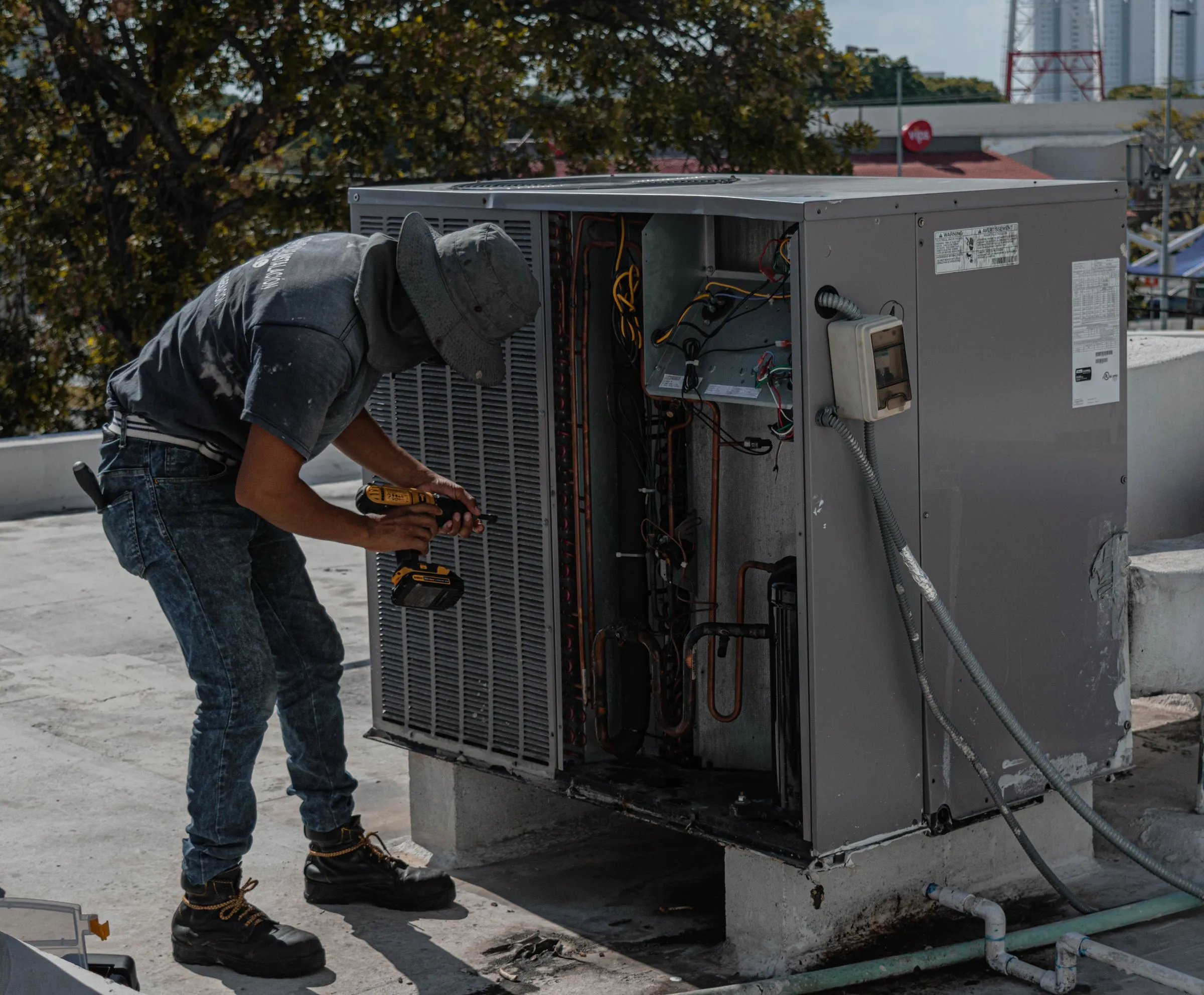 A technician with a cordless drill working on an open air conditioning unit on a rooftop